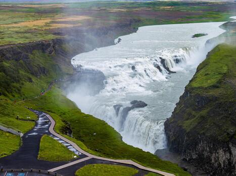 Aerial view shows Gullfoss in southwest Iceland as the Hvita River drops in two tiers into a rugged canyon, with mossy cliffs, mist, paths, and platforms under overcast light. photo