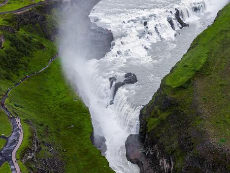 Gullfoss in southwest Iceland shows a stepped cascade turning into a rock cleft. Mist rises as visitors walk on a path and platforms in bright daylight. photo