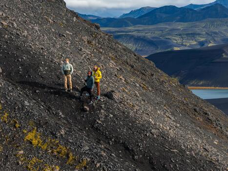 Three hikers stand on a steep volcanic slope with moss patches, a cobalt lake, and layered highlands in Iceland, crisp daylight casting long shadows and sharp textures. photo