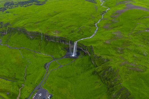 Aerial view shows Seljalandsfoss on Iceland's South Coast, a waterfall dropping from a cliff to a valley, with a river, car park, and paths in soft overcast daylight. photo