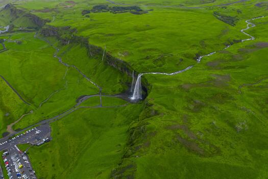 Aerial view shows Seljalandsfoss plunging from a mossy cliff into a small pool in southern Iceland, with a river, parking area, footpaths, and diffuse overcast light. photo