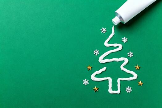 White toothpaste tube squeezing creamy line forming Christmas tree shape on solid green background with tiny snowflakes and golden stars, minimalist New Year greeting card with copyspace for text, fes photo