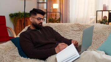 Indian man at home writes carefully in notebook while reading laptop screen, focused on learning. Arabian guy on sofa works calmly preparing for online class with attention, effort and full dedication video