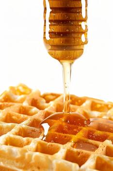Sweet and sticky honey cascades down from a dipper, enhancing the golden texture of a freshly cooked waffle on a plain backdrop photo