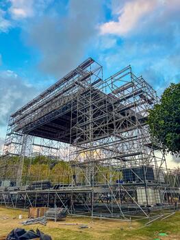 A large metal scaffolding structure is set up in an outdoor area. The sky is partly cloudy, and green trees are visible in the background. photo