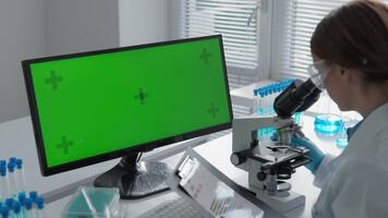 i en modern labb, en forskare i en vit täcka undersöker prover under en mikroskop, använder sig av en grön skärm övervaka för data. detta slingor de allra senaste atmosfär av nuvarande forskning laboratorier video