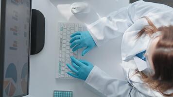 un enfocado investigador en un laboratorio Saco y guantes es mecanografía en un teclado, analizando datos desplegado en un monitor. el escena capturas el esencia de científico investigación y datos análisis. video