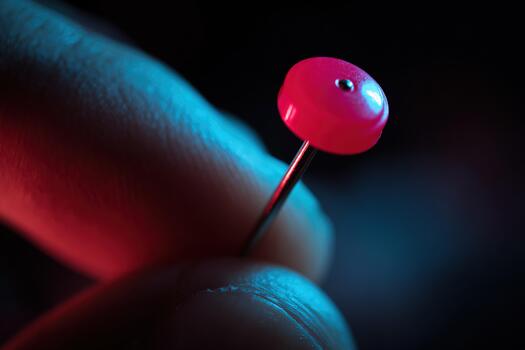 Macro close-up of red push pin held between fingers under colorful lighting photo