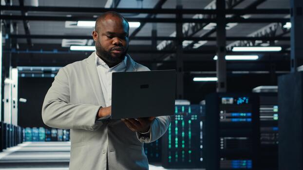 Programmer in data center using laptop, testing equipment failover scenarios. Server farm IT professional uses software on notebook, ensuring data accessibility, camera B photo