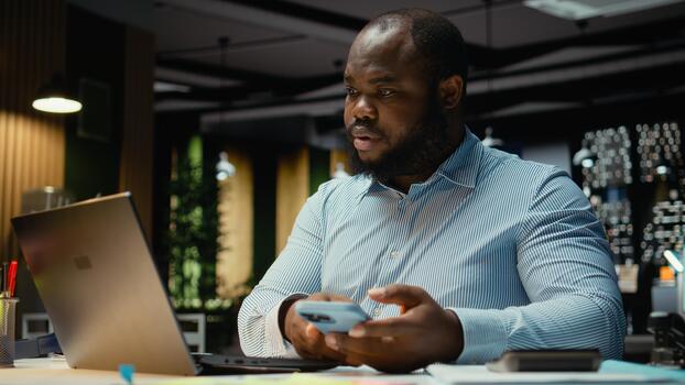 Black office employee checking a notification connected on socials while he does overtime at midnight, checking smartphone at work after hours. Young man reading text messages. photo