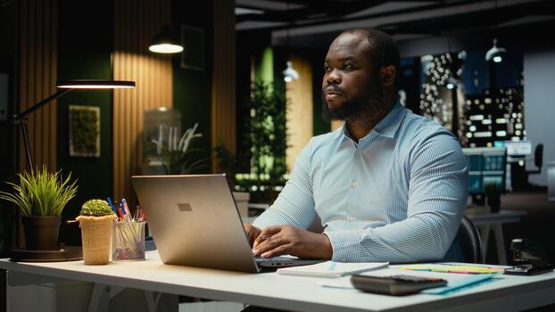 African american guy typing information on laptop and checking reports, proofreading and reviewing data for a new business objective or target. Company management after hours. photo