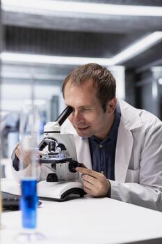 Male scientist evaluating specimen under microscope in clinical lab, using magnifying glass and lens system for a scientific experiment. Medical research and discovery process for bioengineering. photo
