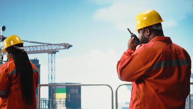 Drilling rig worker using handheld radio device to monitor systems during operations. African american man on offshore platform deck using walkie talkie, calibrating equipment, photo