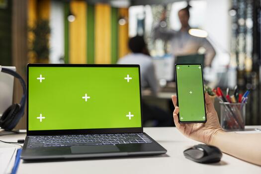 Laptop and cellphone with green screen display in modern office. Closeup of caucasian man using smartphone and laptop with blank chroma key template in workspace of financial firm. photo