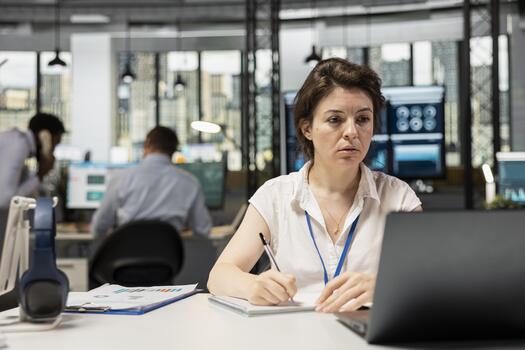 Caucasian female corporate worker writes numbers and company objectives on notepad while reviewing presentation on laptop. Woman analyst takes notes on desk and reading online data on device. photo