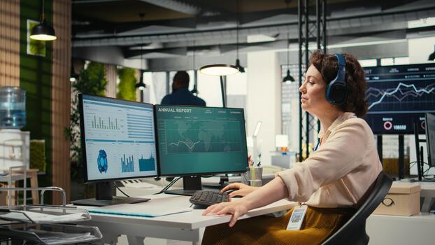 Young businesswoman focused on a podcast while she resolves office tasks, monitoring insights or KPI metrics on pc. Strategy reports for expansion plans with digital tools. photo