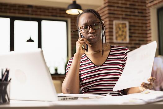 Confused remote worker sits at cluttered table, frustrated by incomplete paperwork while handling business issues via smartphone. Businesswoman visibly upset during tense conversation with colleague. photo