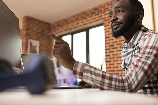 Male remote worker developing software solution using multiple device screens. Black freelancer using laptop and desktop monitor, working with advanced programming languages in brick wall apartment. photo