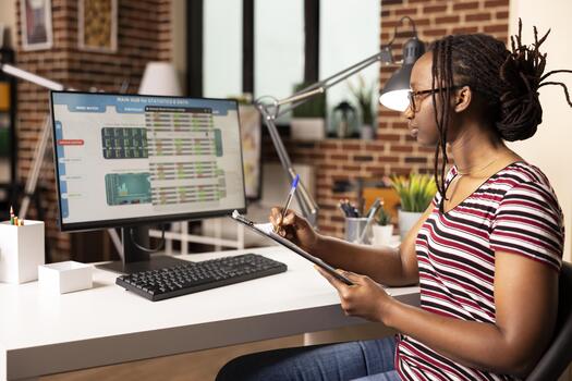 Self employed woman analyzing business charts on desktop pc, comparing metrics with notes from clipboard. Black female manager reviewing financial graphs on computer screen at home. photo