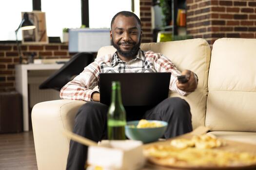 Man seated on sofa with laptop on lap, increasing TV volume using remote control. Smiling male individual switches channels while working remotely, balancing entertainment and project tasks at home. photo