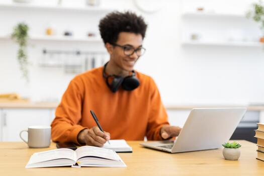 A young man with glasses is working on a laptop while taking notes in a notebook, with a book open on the table. photo