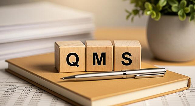 Wooden blocks displaying QMS on a tan notebook with a pen in a bright office setting symbolizing quality management system implementation and business process optimization for enhanced efficiency photo