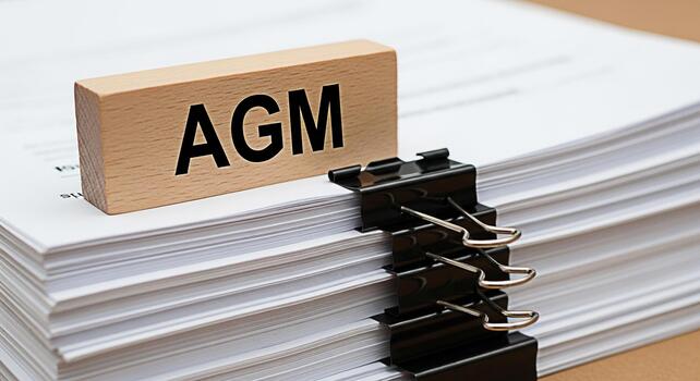 Wooden block displaying AGM sitting atop a stack of white papers secured with a binder clip symbolizing the importance of the Annual General Meeting in a corporate setting conveying organization and f photo