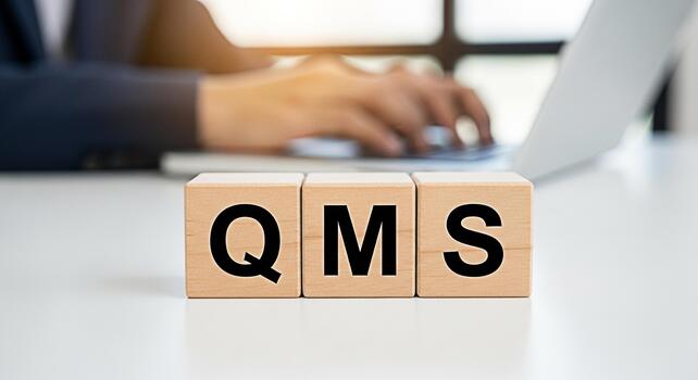 Business professional typing on a laptop in a bright office with wooden blocks spelling QMS representing Quality Management System and a commitment to high standards and continuous improvement photo