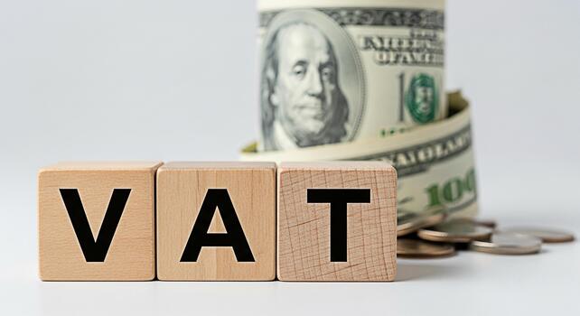 Wooden blocks spelling VAT stand prominently in a clean studio setting symbolizing valueadded tax with a roll of US currency and coins representing financial implications and economic concepts photo