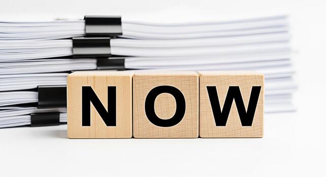 Wooden blocks spelling NOW stand prominently in front of a large stack of paperwork on a clean white surface symbolizing urgency action and the importance of immediate attention to tasks and deadlines photo