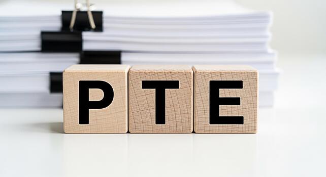 Wooden blocks spelling PTE on a white desk with a stack of papers in the background representing the Pearson Test of English academic exam preparation and success in a bright and clean office environm photo