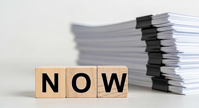 Wooden blocks spelling NOW sitting on a white desk next to a large stack of documents emphasizing urgency and the importance of immediate action in a busy office environment photo