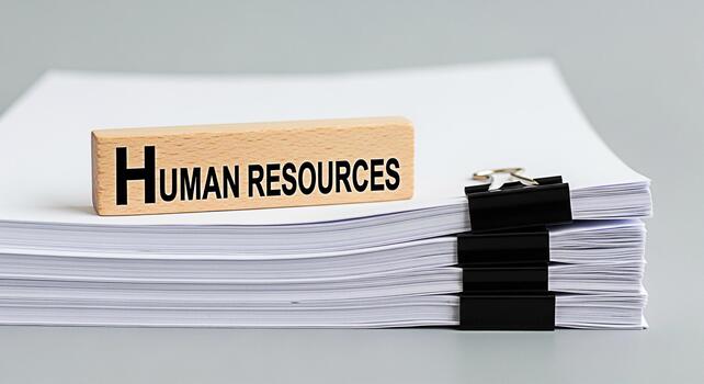 Wooden block displaying Human Resources rests atop a stack of white papers secured with binder clips in a brightly lit office symbolizing organization and efficient HR management photo