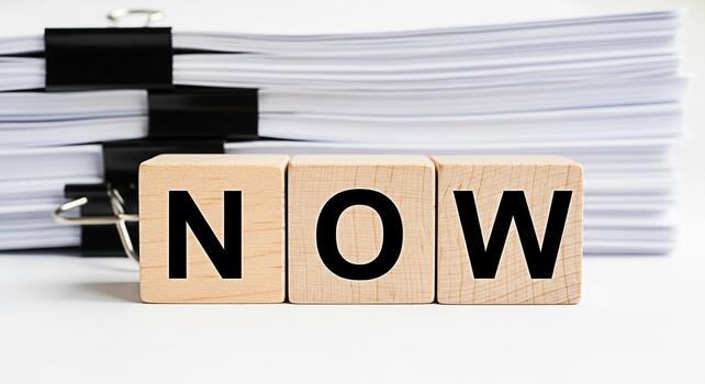 Wooden blocks spelling NOW in front of a stack of papers emphasizing urgency and immediate action in a busy office environment conveying a sense of focus and productivity for time management and effic photo