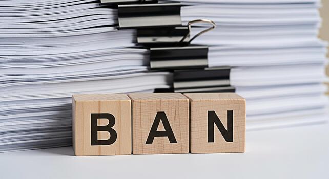 Wooden blocks spelling BAN stand defiantly against a towering stack of documents on a clean white surface symbolizing restriction and the urgent need to control excessive paperwork in a modern office photo