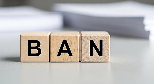 Wooden blocks spelling out BAN on a neutral surface symbolizing restriction and prohibition in a clean minimalist setting conveying a message of censorship and control with a stack of paper in the bac photo