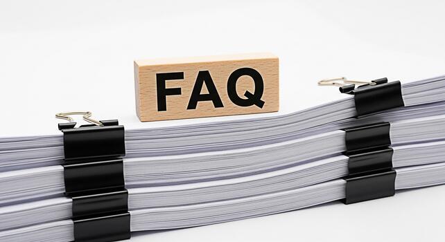 Wooden block displaying FAQ rests on a stack of paperwork secured with binder clips in a bright clean office environment representing readily available information and customer support photo