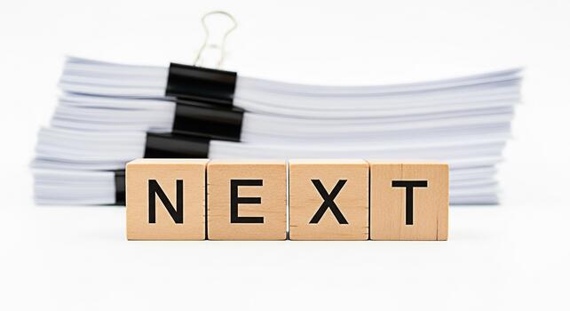 Wooden blocks spelling NEXT in front of a stack of papers on a white surface representing forward progress future planning and a positive outlook for business and personal growth photo