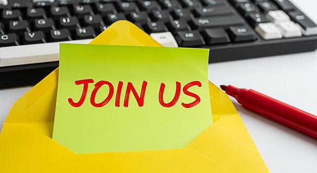 Bright yellow envelope on a desk with a green sticky note reading Join Us next to a keyboard and red marker inviting participation and creating a welcoming and collaborative atmosphere photo