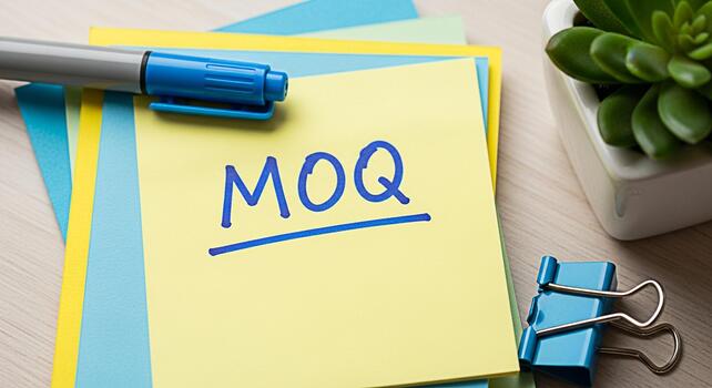 Closeup of a yellow sticky note displaying MOQ on a bright wooden desk surrounded by colorful paper a pen and a succulent representing minimum order quantity and business planning photo