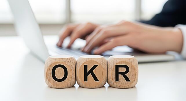 Hands typing on a laptop with wooden blocks displaying OKR on a white desk symbolizing Objectives and Key Results in a bright office environment representing business strategy and goal setting photo