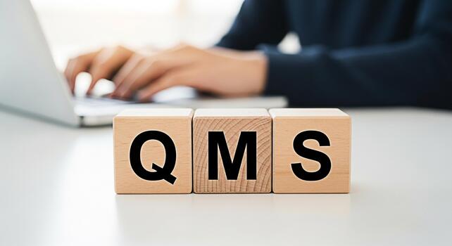 Hands typing on a laptop with wooden blocks spelling QMS on a white desk representing quality management system implementation in a modern office conveying efficiency and systematic approach photo