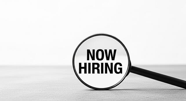 Magnifying glass focusing on Now Hiring text on a gray surface symbolizing job opportunities and recruitment efforts in a minimalist and professional setting conveying a message of active hiring and c photo