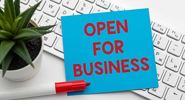 Bright blue note stating Open for Business placed on a white computer keyboard with a red marker and a small potted plant conveying a welcoming and readytoserve business environment photo