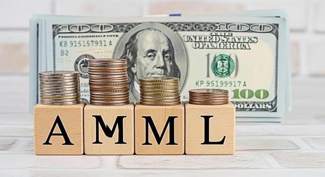 Stacks of coins resting on wooden blocks spelling AML AntiMoney Laundering in front of a stack of hundred dollar bills on a white brick background representing financial compliance and regulatory over photo