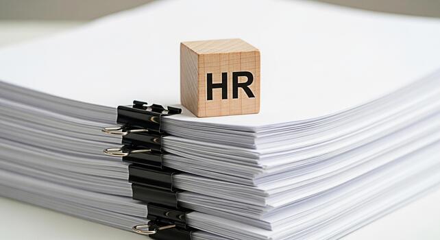 Wooden block with HR on top of a stack of white papers secured with binder clips in a bright office setting representing human resources management and administrative paperwork photo