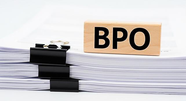 Wooden block displaying BPO resting atop a stack of documents secured with binder clips on a clean white surface symbolizing business process outsourcing and efficient document management photo