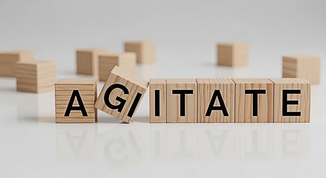 Wooden blocks spelling Agitate on a white surface with one block tilted creating a sense of disruption and unease symbolizing the act of stirring up emotions or causing unrest in a minimalist setting photo