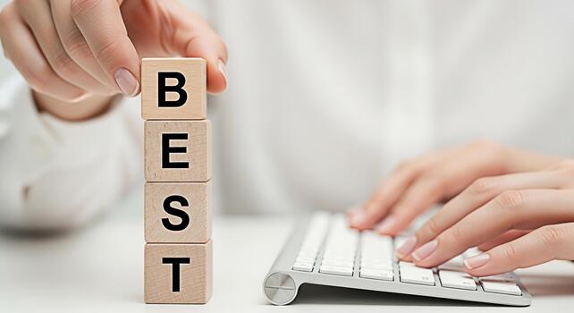 Focused hands stacking wooden blocks spelling BEST next to a keyboard on a clean white desk symbolizing achievement and success in a professional and minimalist working environment photo