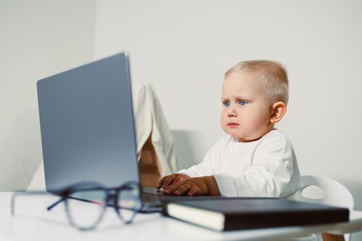 Curious toddler explores technology while seated at a desk displaying focus and wonder in a well-lit room photo